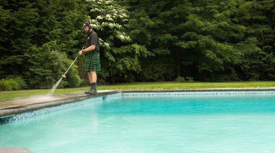 Man in a kilt pressure washing the poolside patio next to a clear blue swimming pool surrounded by green trees.