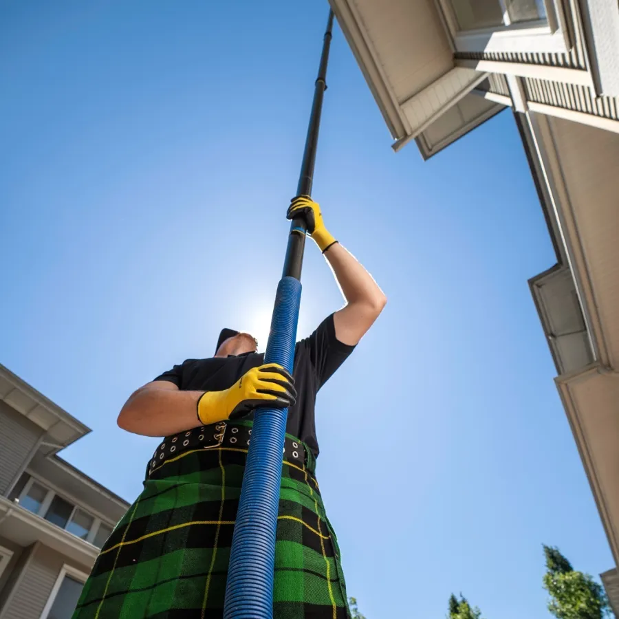 Person wearing a green plaid kilt and yellow gloves using a long blue pole for gutter cleaning on a sunny day.