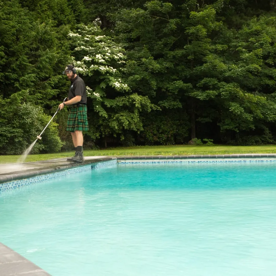 Man in a kilt pressure washing the stone deck around a backyard swimming pool surrounded by green trees.