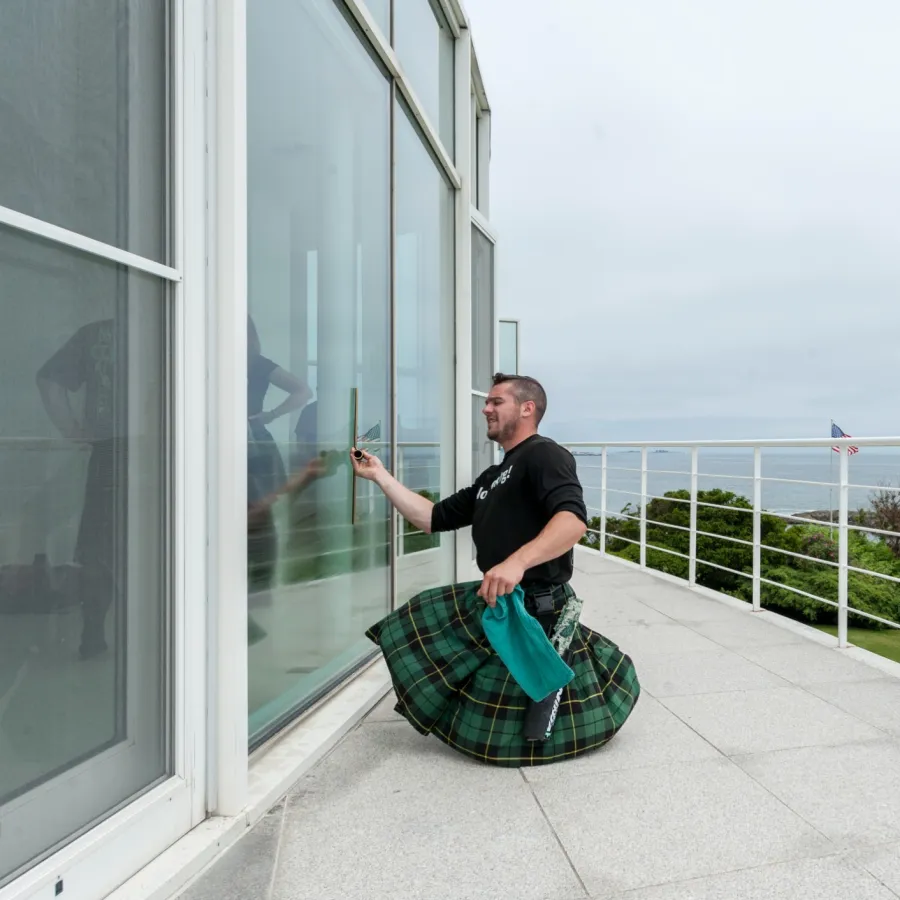 Man in a green plaid kilt cleaning a large glass window outside on a terrace overlooking the ocean