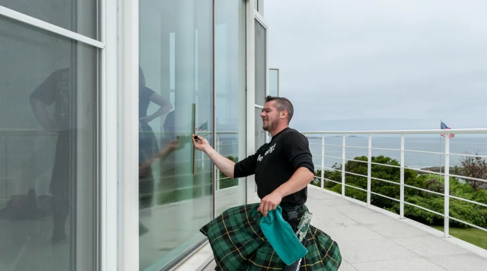Man in a green plaid kilt cleaning a large glass window outside on a terrace overlooking the ocean