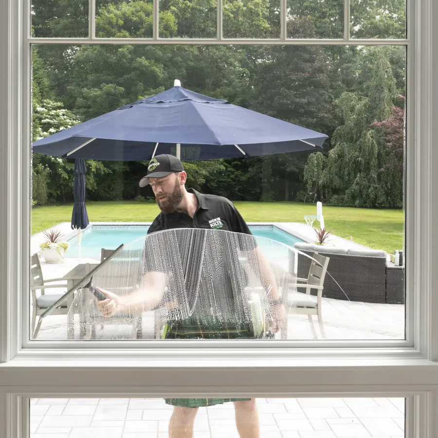Man cleaning a large window with a squeegee outside by a pool under a blue umbrella on a sunny day
