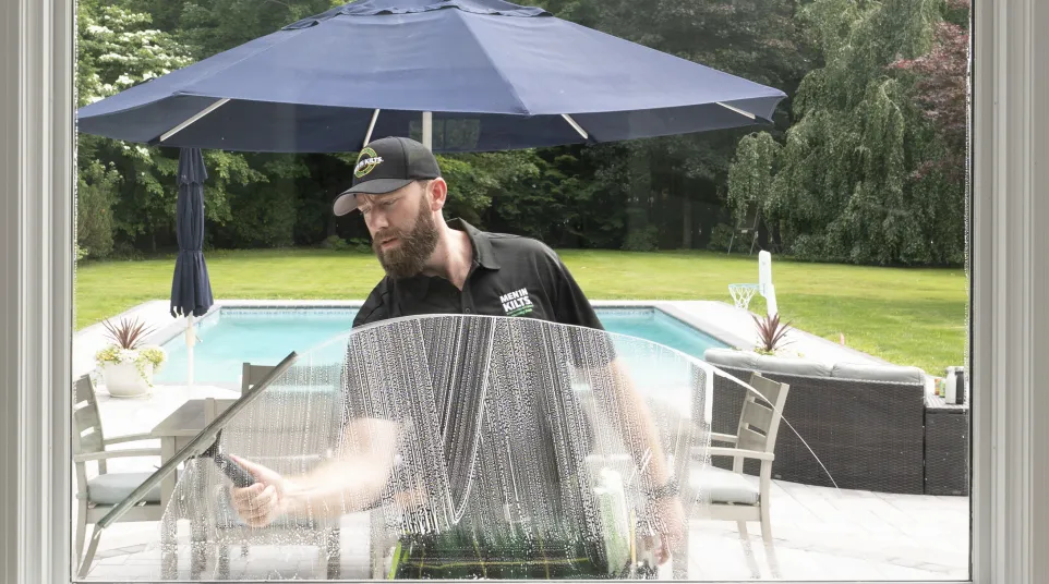 Man cleaning large window outside with squeegee near pool and patio furniture under blue umbrella