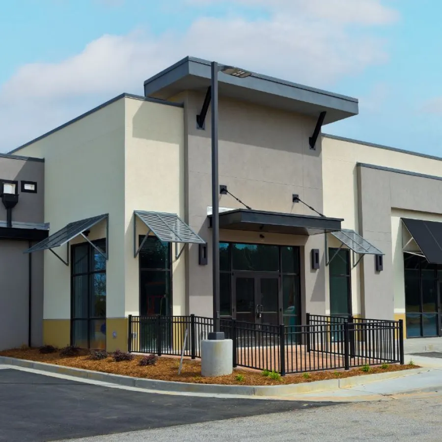 Modern commercial building with beige and gray walls, large windows, awnings, and a small fenced patio area.