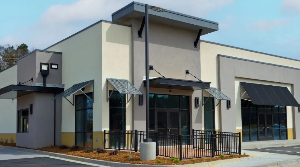 Modern commercial building with large windows, metal awnings, and a fenced patio area under a blue sky.