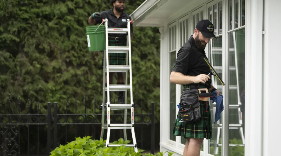 Two workers in kilts cleaning windows of a white house surrounded by greenery using professional tools.