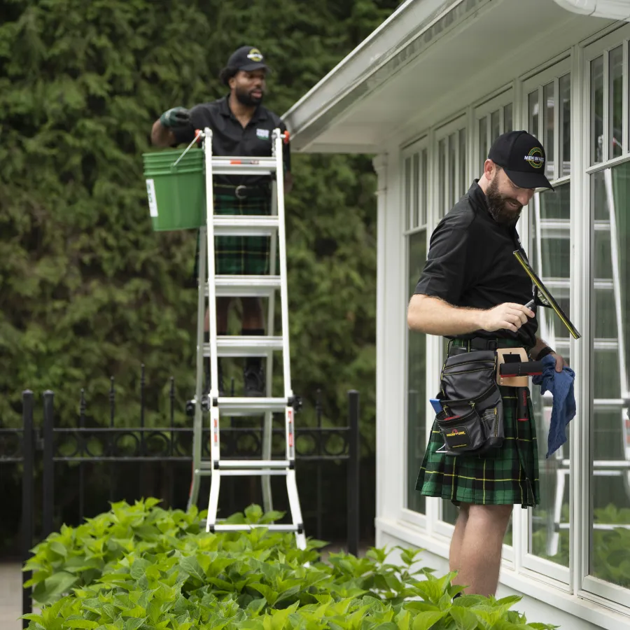 Two men wearing kilts cleaning windows and gutters on a white house surrounded by greenery