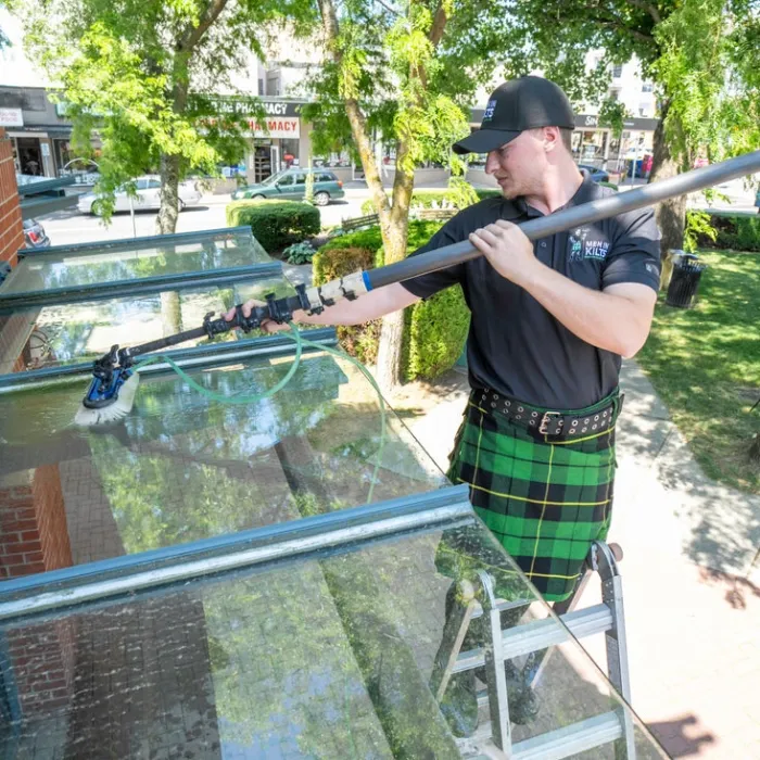 A man in a kilt cleans windows using a long pole on a sunny day.